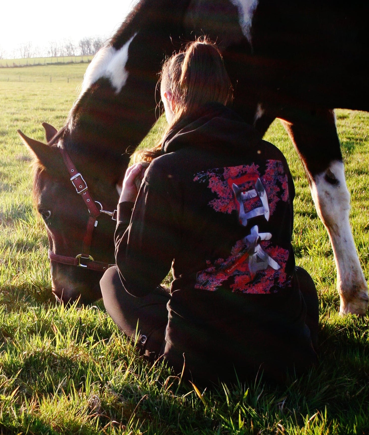 Photo de cheval broutant avec sa cavalière vue de dos assise à côté de lui. La cavalière porte un sweat noir et rouge sur le thème du cheval et du Japon, avec la tête du cheval inscrite dans des kanji japonais.