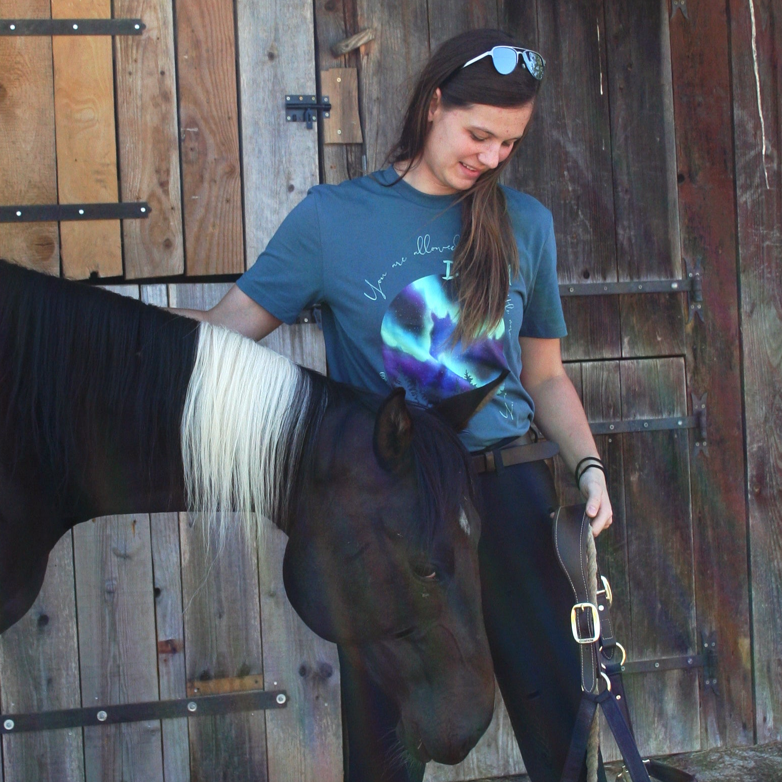 Cheval avec sa cavalière qui porte un t-shirt au motif d'aurores boréales qui dessinent un cheval dans le ciel.